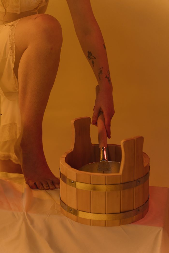 our-journey-01 A woman kneels, holding a ladle beside a wooden sauna bucket, creating a warm and serene atmosphere.